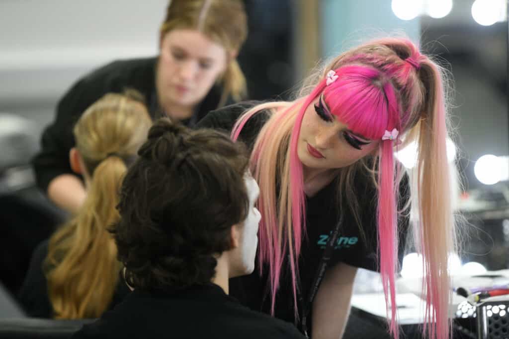 Female theatrical makeup learner with pink hair applies make up to another learner